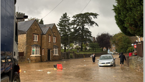 A picture of a road in Lavendon that has been hit by heavy flooding. A car can be seen partially submerged as people wade through the water. 