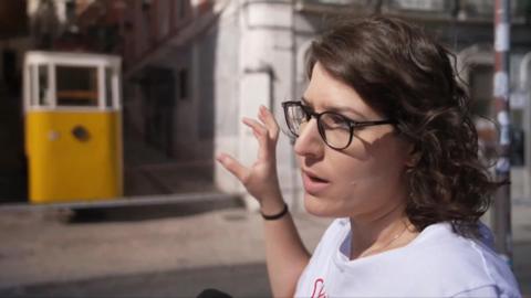 Rasha Abdo wearing a white top and black rimmed glasses gestures towards the undamaged yellow and white funicular carriage.
