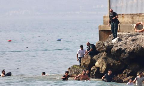 Men swimming as police officer standing on rocks holds arm of man in white t-shirt