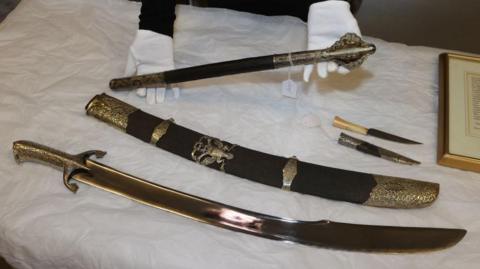 Old weapons laid out on a table - a scimitar (sword/sabre) and scabbard, a mace, and a dagger and sheath.
