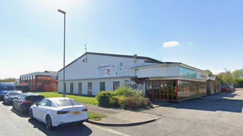 A one-storey white building has the words The Future Building on the side with brown folding doors at the entrance and brown framed windows. Beyond the building is another industrial building