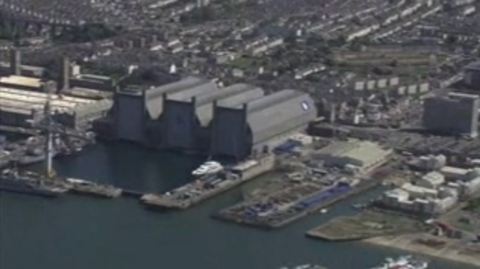 A birds eye view of the Devonport dockyard. There are large buildings and houses. The sea is a dark blue. 