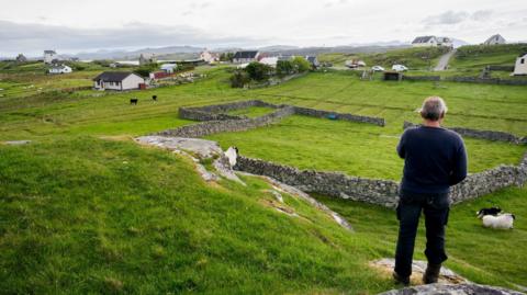 Croitear a' coimhead air an talamh aige le caoraich agus cu chaorach 