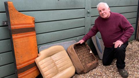 Mark Fishpool is wearing a burgundy jumper and black trousers, while kneeling on the ground next to several broken leather car seats.