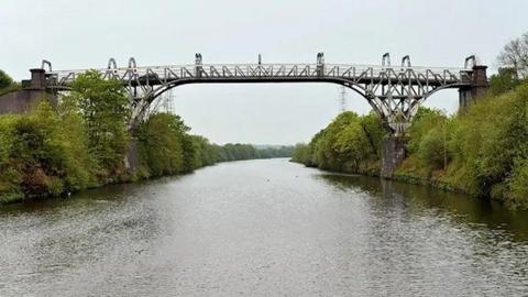A metal bridge straddles two wooded banks over a large river on a clear day.