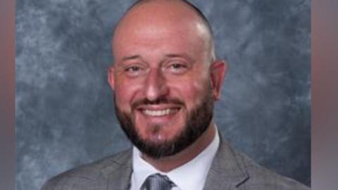 Karl Perks, a bald man with a beard wearing a grey suit with white shirt and grey tie. He is positioned against a dark grey backdrop and is smiling for the photograph.