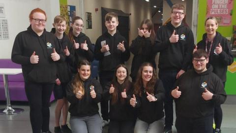 A dozen young people, all wearing black hoodies and holding their thumbs up, smile in a posed group photograph.