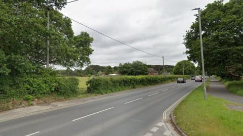 A Google Street View of an A road. A hedge runs alongside the left of the road, with a field beyond. On the right is a footpath.