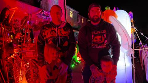 Two men, both in Halloween jumpers, with two young children in front of spooky inflatables and props.