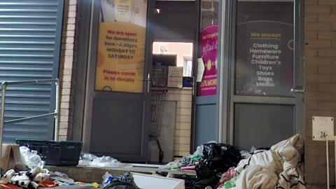 Bags and boxes lie in a disorganised way in front of a set of a doors. There is signage on the doors indicating it is a charity shop with opening hours and the type of items accepted.