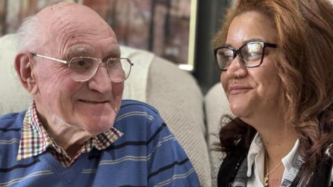 91-year-old Don Way and his carer Jamaica Bell are exchanging a knowing glance after Don teased her for forgetting to put a sweetener in his coffee. Don's wearing a smart blue V-Neck jumper with stripes on it over a checked shirt. Sitting to his left is Jamaica. Also wearing glasses, her long hair falls over the shoulders of the black and white cardigan she's wearing over a white shirt. 