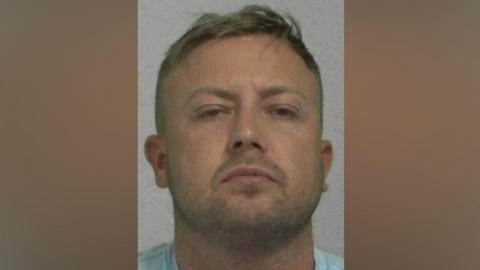 Mugshot of Watson. He has faint stubble and short blond hair.