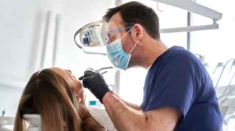 A dentist wearing a visor and a mask with dental tools and examining a woman's mouth