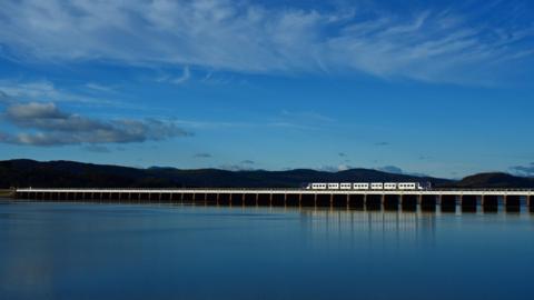A Northern train on the Furness line crossing the bay at dusk, with the fells in the background. The water and sky appear as a deep blue. The five or six carriage train - mainly white but with blue detailing - is on an extremely long and low viaduct which ploughs a straight line across the wide expanse of water.