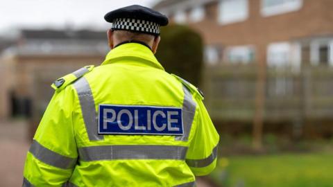 A police officer wearing a hi-vis jacket walks around what appears to be a housing estate.