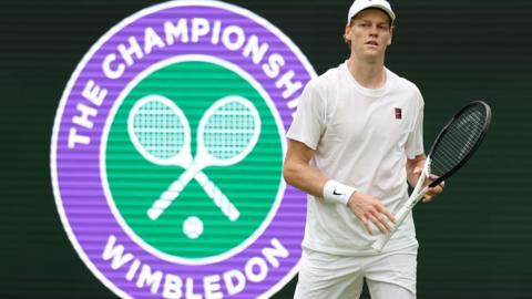 Jannik Sinner holds his tennis racquet while standing in front of a large purple and green logo that reads The Championships Wimbledon