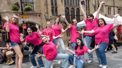 A group of nine people are outside Market Hall in Durham with their arms in the air and joyous looks on their faces. They are all wearing pink t-shirts, which say "Durham Fringe Festival" on the front.
