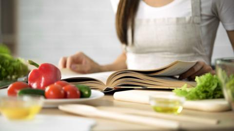 A woman reading a cook book with fresh vegetables and kitchen tools on table
