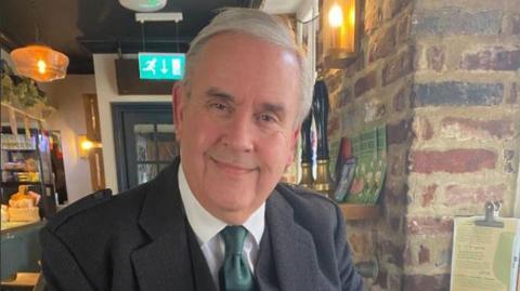 Dave smiles at the camera - he is wearing a dark coat over a suit and a green tie. He is sitting in a pub, with exposed brick wall to the right.