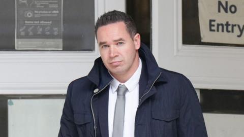 A man with short black hair wearing a large dark blue coat. He is looking into and walking towards the camera.