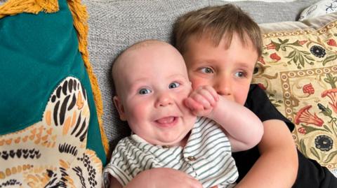 An older child sits on a couch holding a smiling baby in a striped outfit, both nestled among patterned cushions.