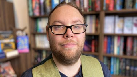 Alistair smiling at the camera wearing a beard, waistcoat and glasses standing in front of bookshelves