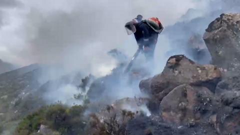 Firefighter dampening flames among rocks and grassland as plumes of smoke rise into the air