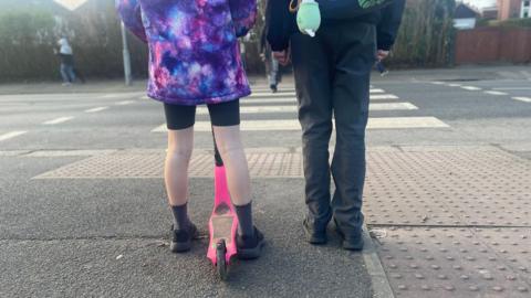 The back legs of two school children, one with a scooter between their legs, standing at the side of the road with a zebra crossing in front of them.