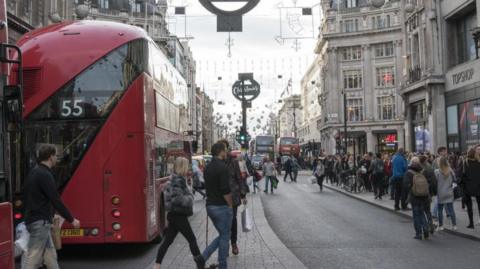 Shoppers crossing Oxford Street at Christmas time including some people weaving between two buses, and a crowded pavement on the other side of the road