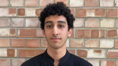 A young man with a black shirt looks at the camera and stands against a brick wall