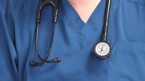 A closeup of the neck and shoulders of a nurse wearing v-neck blue scrubs with a black stethoscope hanging around the neck.