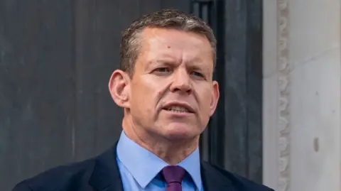 A man, Rhun ap Iorwerth, is in a navy suit.