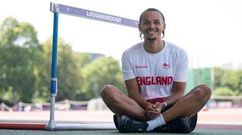 A smiling Sam Bennett wearing a Team England team shirt, sitting next to a hurdle