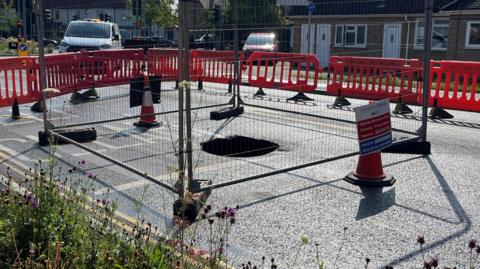 A sinkhole is in the middle of a road which has been closed. Around the sinkhole is a metal fence and cones with signs. Surrounding that are red barriers which have closed the road.