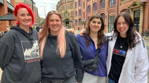 Four women standing together as they head off to Leeds festival, one has red hair and another has pink hair, and two have brown hair. 