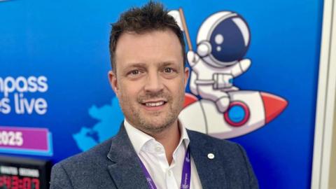 A man in a suit and white shirt smiles as he stands in front of a blue background, with a cartoon drawing of a space rocket on it. He has short dark hair, and is wearing a lanyard.
