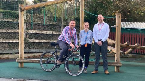Two male and one female head teachers in a school playground. One of the men is sitting on a bicycle.
