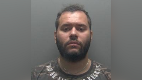 Custody photo of Daniel Barbosa against a grey background. He has short dark hair and facial hair. He is wearing a gold chain and camouflage style top with writing around the neck. 