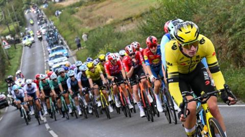Tens of cyclists race up a hill. The lead cyclist wears a yellow jersey, helmet and rides a yellow and black bike. 