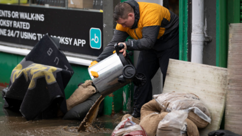 Business owner stood in shop opening, poruing out a binfull of water into a flooded street which is full of muddy water, sandbags and rubbish