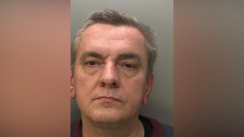 A police mugshot of a man with grey hair. He has brown eyes and is clean shaven.