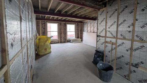 The interior of a building with a window on the far wall and wooden frames with plastic sheeting stretched between them on all walls. The wooden beams of the ceiling are exposed