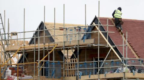Picture of two new homes have roofing tiles put on. There is scaffolding and a worker is on the roof of the one of the houses. 