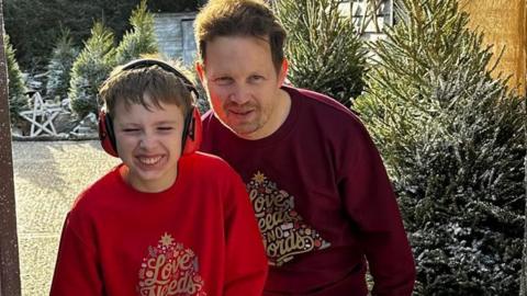 Tommy, wearing ear defenders and a red christmas jumper smiles, next to his dad James, who is also smiling and wearing a burgundy christmas jumper