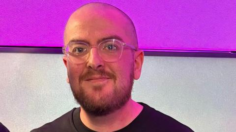 A man wearing glasses and a black jumper smiles as he stands in a studio with a purple and grey wall behind him.