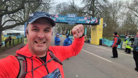 Stuart Byrne taking a selfie at the finish line of the Cambridge Half Marathon. He is smiling at the camera and holding up a fist. He is wearing a blue cap and bright red jersey. Behind him is a finish line over a stretch of road. 