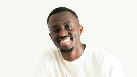 Joseph Charm is smiling at the camera while wearing a white crew neck top. He has dark hair and facial hair. He is in front of a white background.