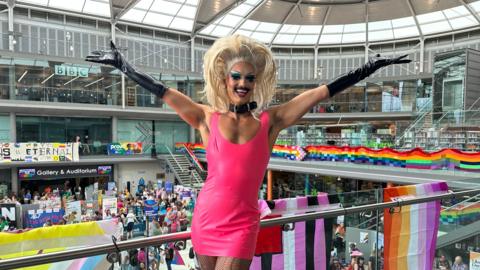 Crystal is standing in the middle of the image wearing a pink dress. She has blonde hair and has both arms in the air. Behind her are flags which are hanging down from a balcony and people are walking. 