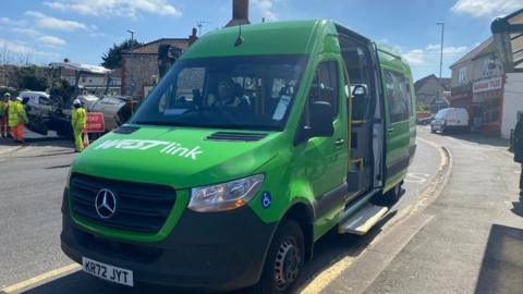 A green WESTlink minibus parked in a bus lane on a sunny day.