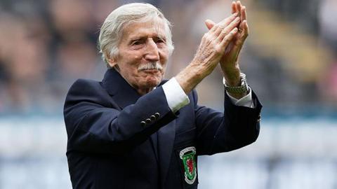 Cliff Jones, who has white short hair and a moustache, has his arms raised clapping. He has a blue blazer on with the Wales logo.